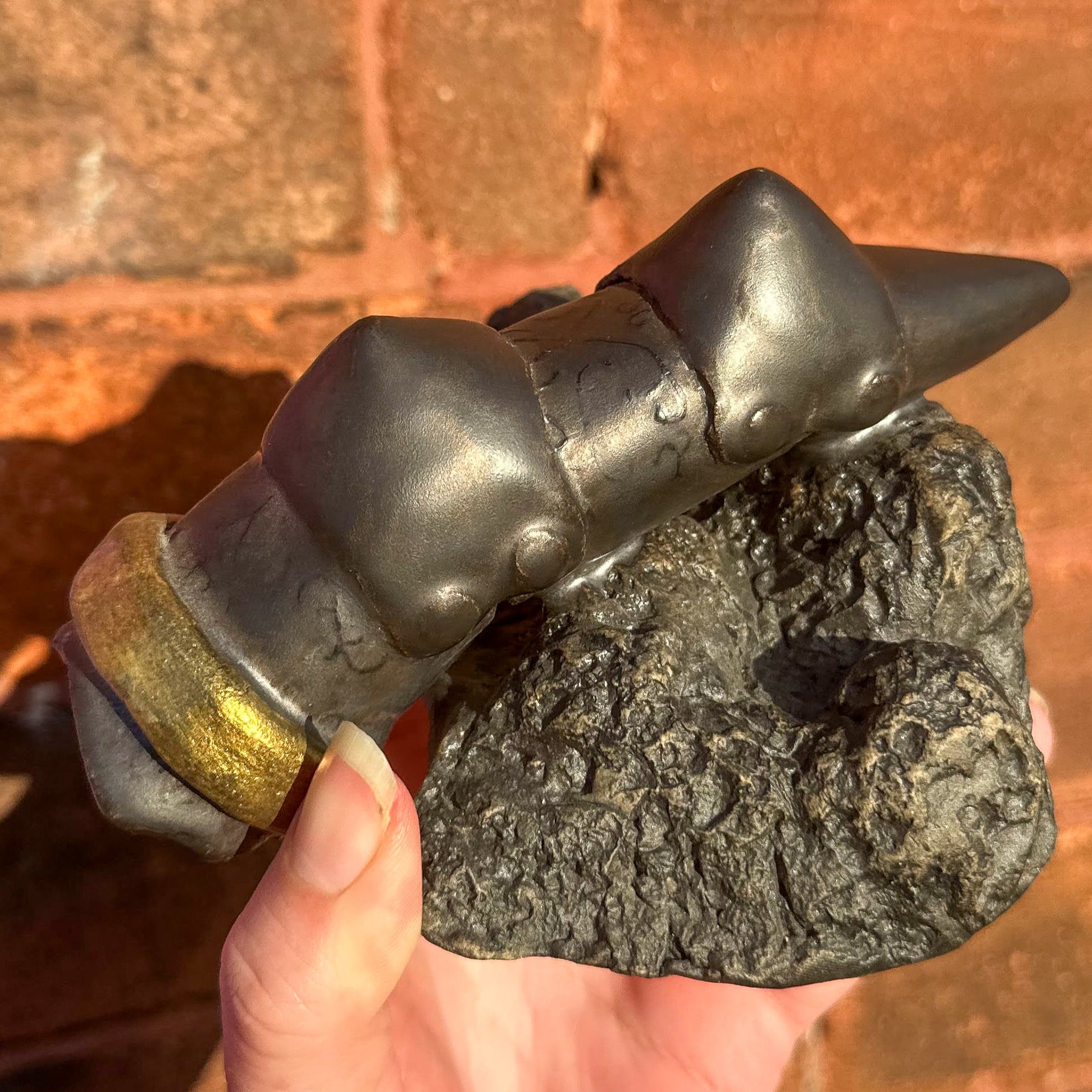 A ceramic sculpture of a severed armoured finger, the amour is a black aged metal finish, with subtle pattern on the finger segments. The two knuckle pieces of the amour are pointed. There is a shiny gold ring round the base of the finger just above the severed edge. The finger is propped slanted upwards on a grey and black rocky base.