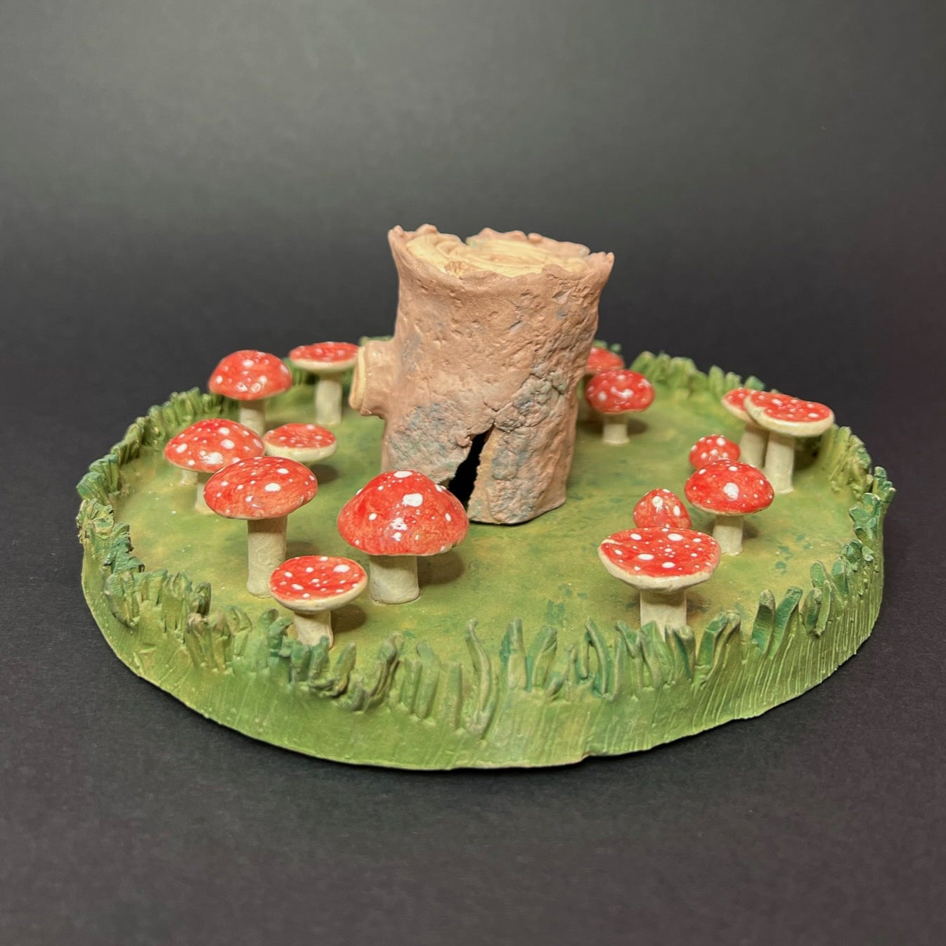 A ceramic sculpture featuring a green base which looks like green grass with a textured grass ring around the outside. Within that is a fairy ring of mushrooms that have glossy red caps with white spots and white stems. At the centre there is a textured wooden log standing up on end.