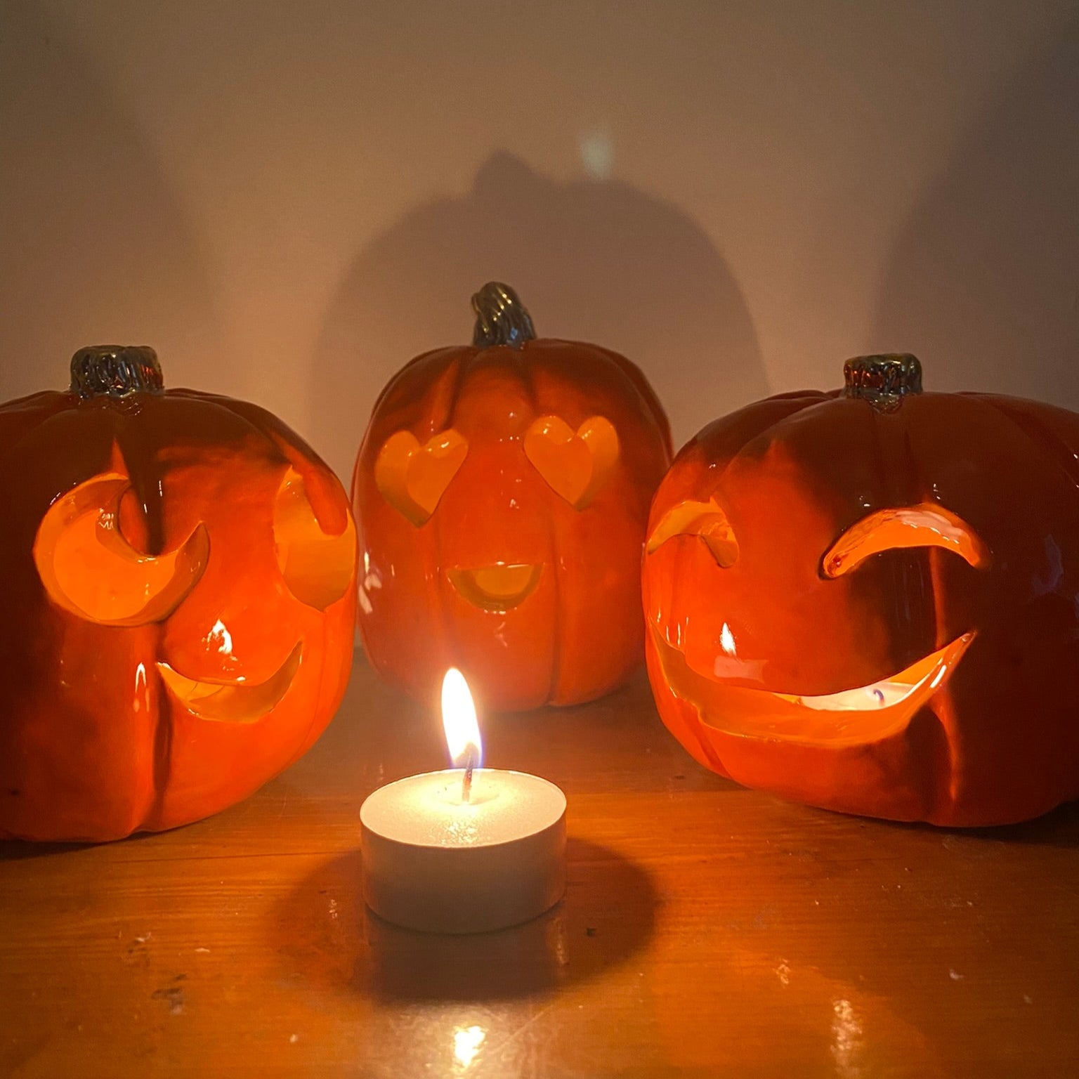 3 ceramic sculptures of pumpkins, they are all bright orange with carved faces and small golden stalks. The first face is looking up and to the right with a small smile. The second has two hearts for eyes and has an open mouthed smile. The third has eyes that are arched slits and a wide smile like it's laughing. In the centre in front of them there is a small tea light, they also each have one lighting them up from inside.