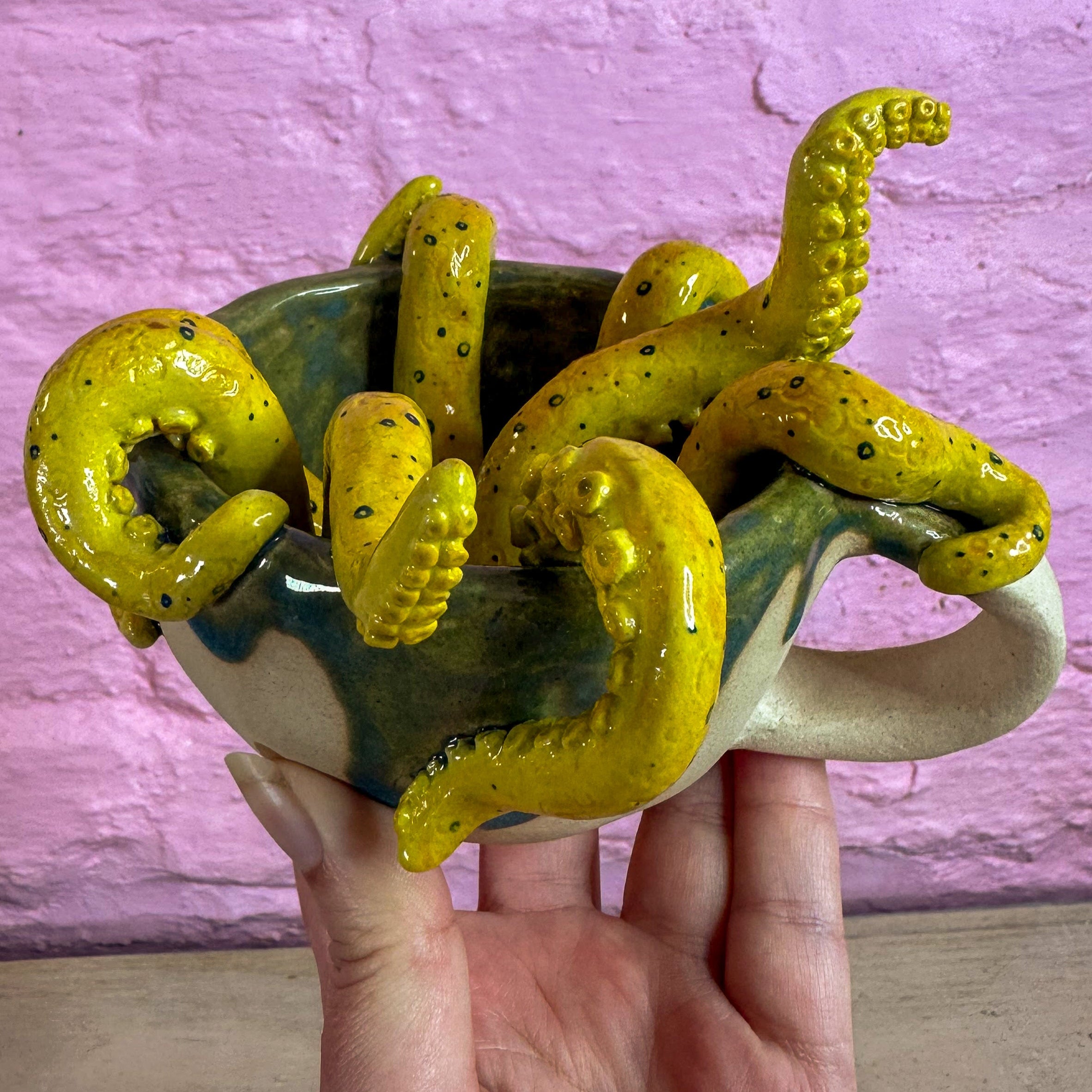 A ceramic sculpture comprising of a group of 8 tentacles which are yellow, with a darker tone over the top and blue rings and small dots. They are reaching and curling out the top of a mug which has green spilling out from the inside and down the top third of the mug.