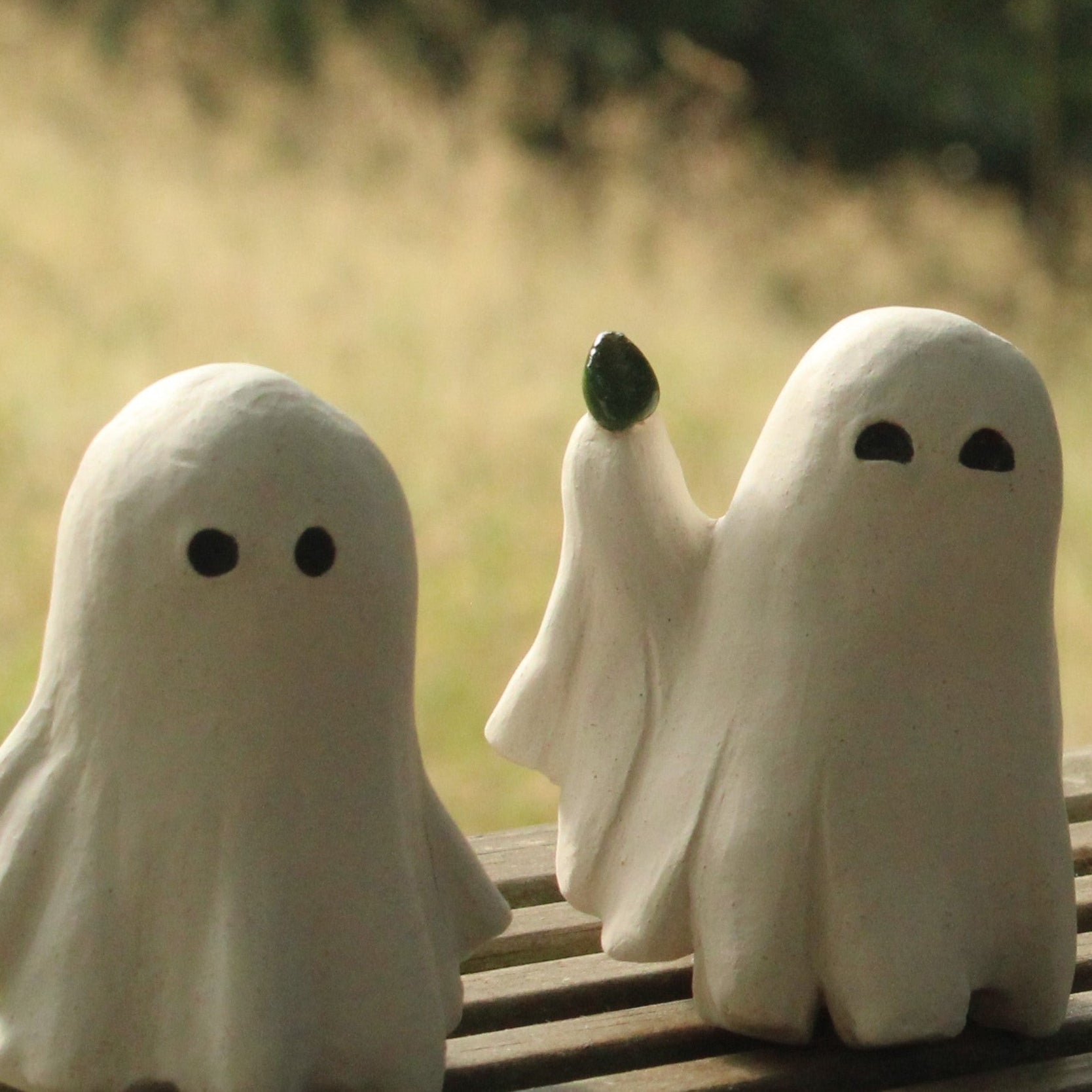 Two ceramic sculptures of ghosts in white sheets, one is just stood upright and the second has one arm in the air holding a single green leaf and it's eyes are arched up like it's smiling.