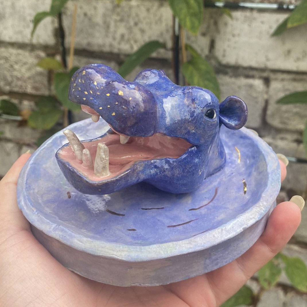 A ceramic shallow blue dish with golden lines for ripples, coming from the centre is a sculpture of a hippos head like it's breaking through the water. It is a purple and blue blended colour with golden freckles across it's nose. It's mouth is open exposing the pink of it's mouth and white teeth.