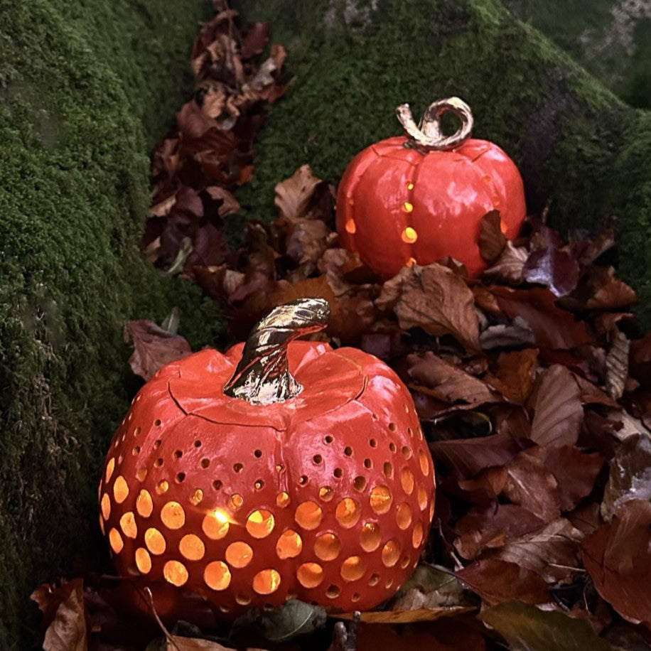 Two ceramic pumpkin sculptures, they both feature circles carved into them in decorative patterns which show candlelight spilling out. The outside are bright orange, the insides are a light yellow/orange and they both have curved golden stems. They are sat amongst fallen leaves at the base of a mossy tree.