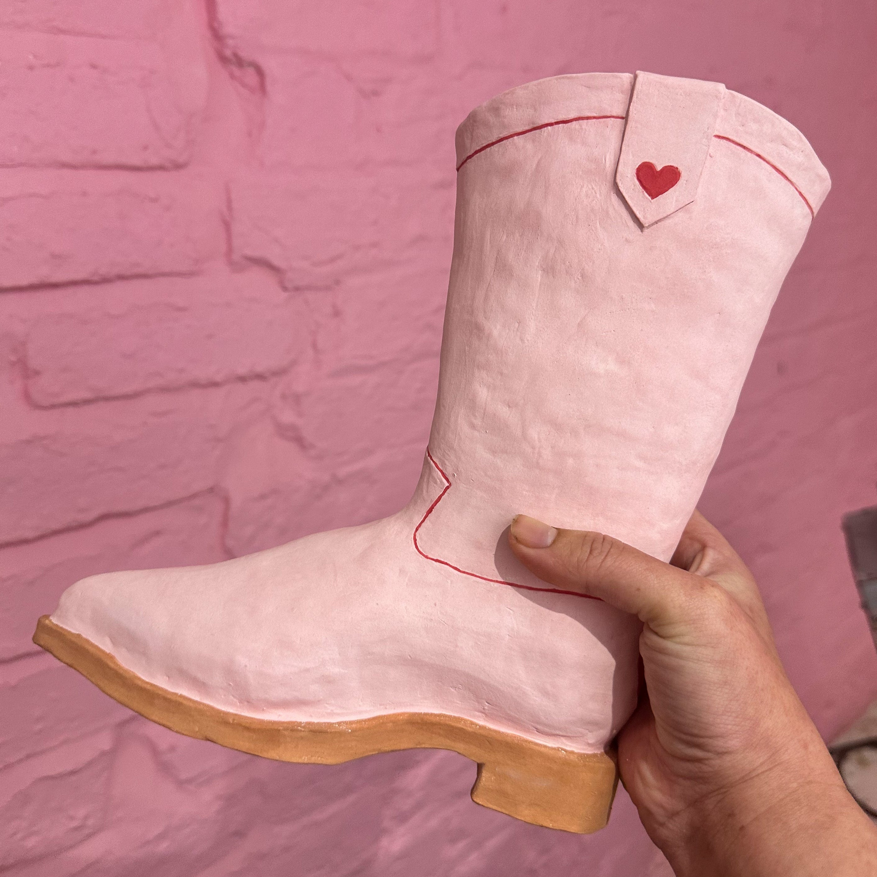 A ceramic sculpture of a pink cowboy boot with a light brown sole and red line details along the seamlines and a heart near the top.