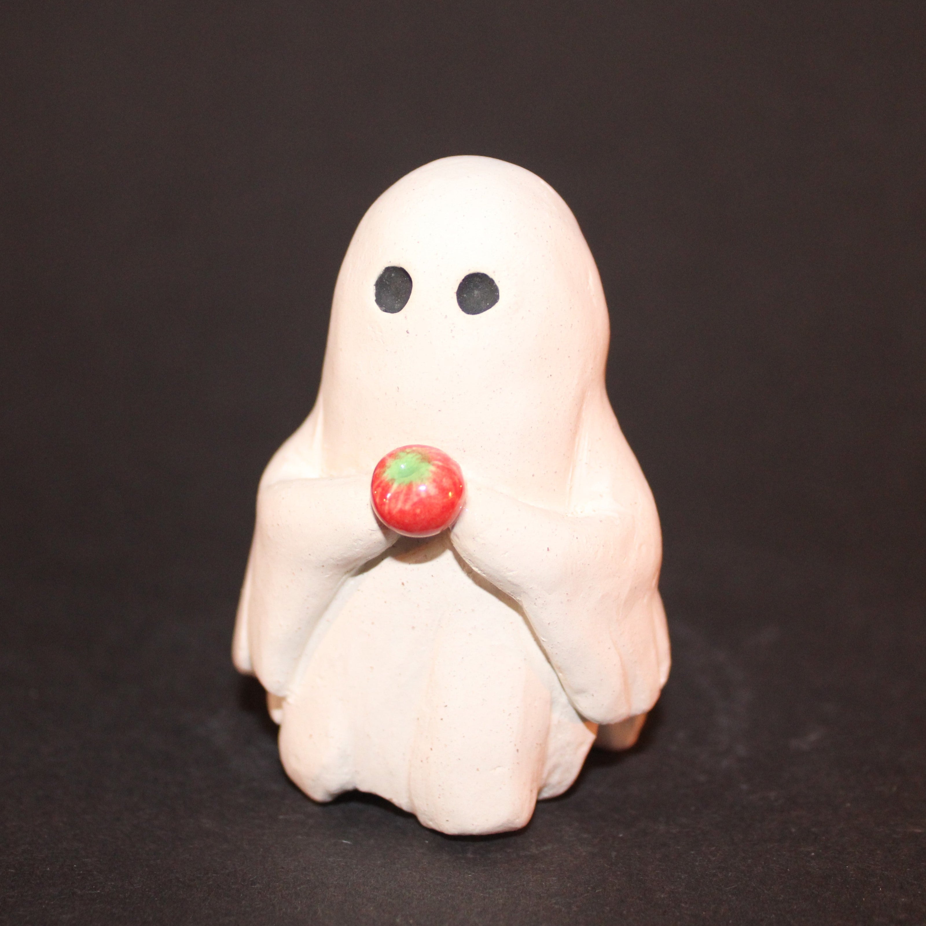 A close up photograph of a ceramic sculpture of a white sheet ghost with it's two arms in front of it holding a small apple that is green at the top and fade into red.